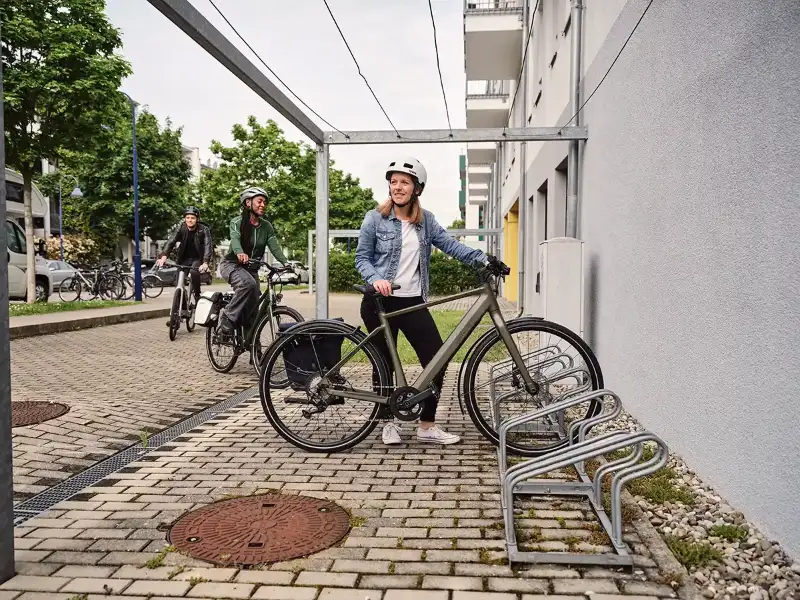 Eine Frau in blauer Jeansjacke parkt ihr grünes E-Bike, eine weitere Frau und ein Mann fahren den Fahrradstellplatz an Eine Frau in blauer Jeansjacke parkt ihr grünes E-Bike, eine weitere Frau und ein Mann fahren den Fahrradstellplatz an